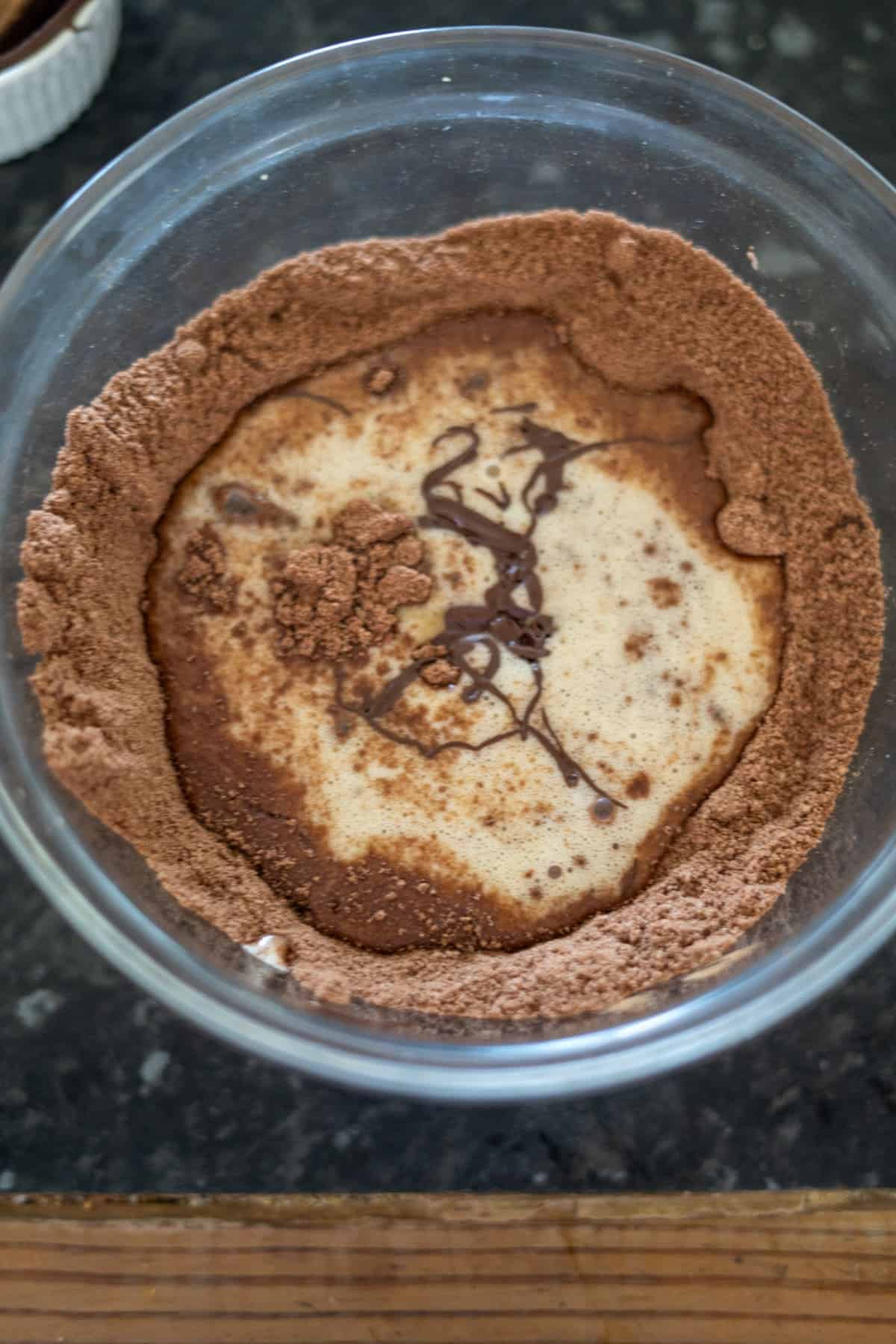 A glass bowl with flour, cocoa powder, and a puddle of liquid ingredients partially mixed on a dark countertop.