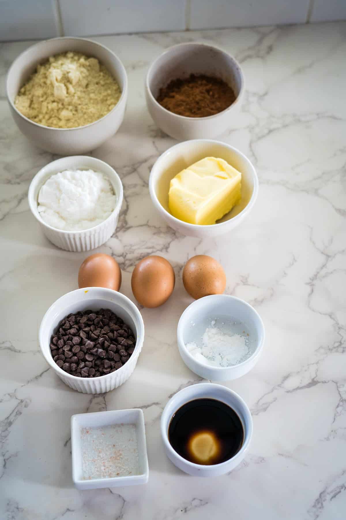 Bowls of flour, cocoa powder, sugar, butter, chocolate chips, salt, vanilla extract, and baking powder are arranged on a marble surface, along with three eggs.