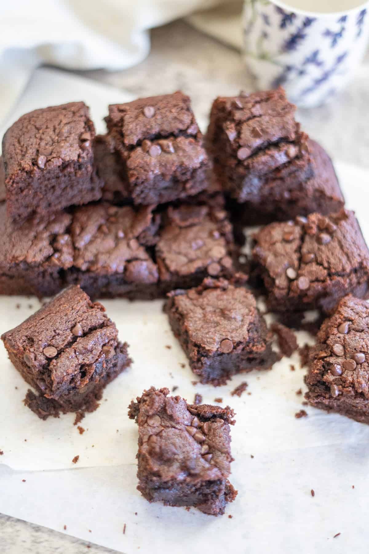 A batch of chocolate brownies cut into squares, stacked on a white parchment paper. Some pieces are scattered around. A partially visible mug is in the background.