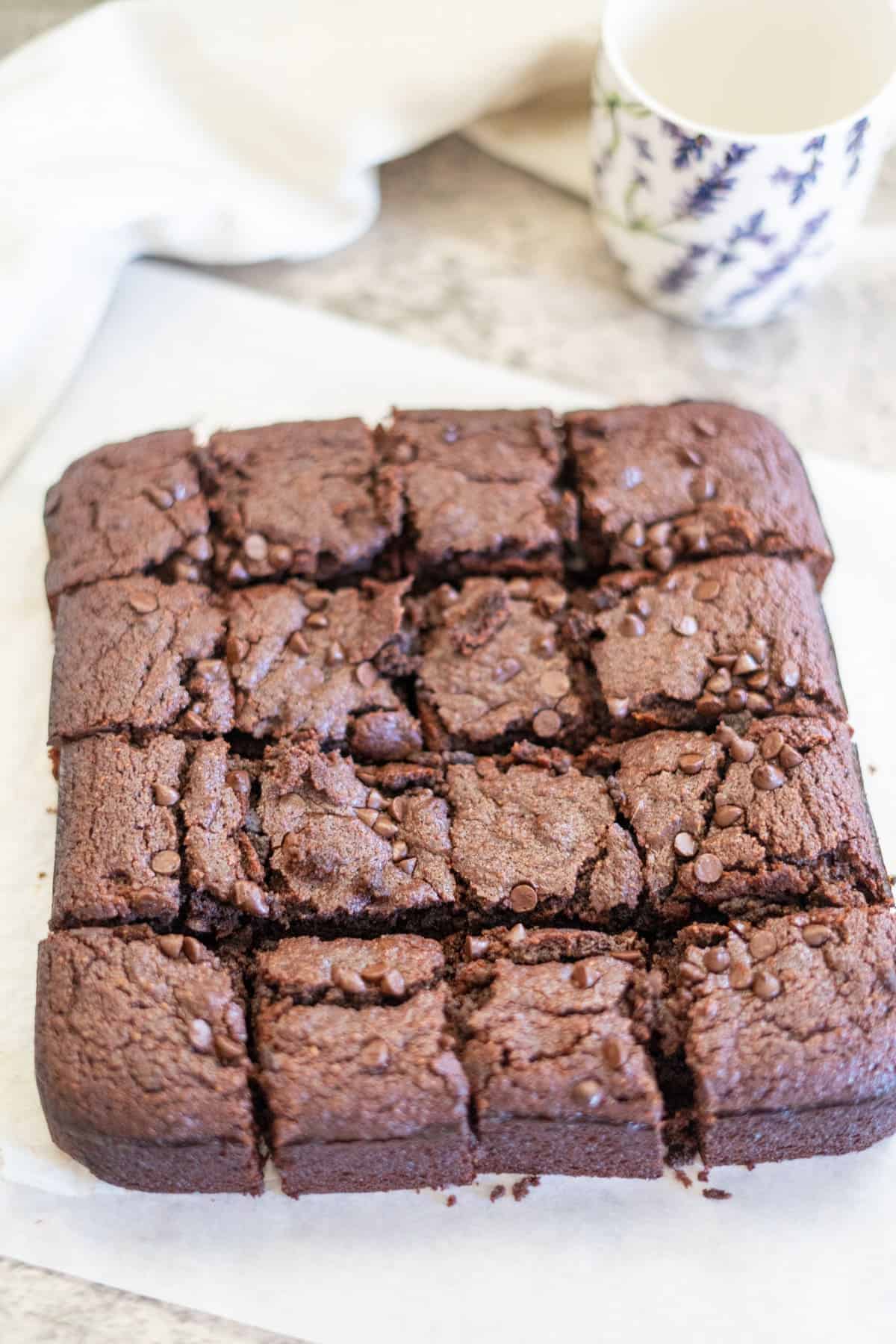 A square chocolate brownie cut into 16 pieces is on a sheet of parchment paper. A white mug with a floral pattern is in the background.