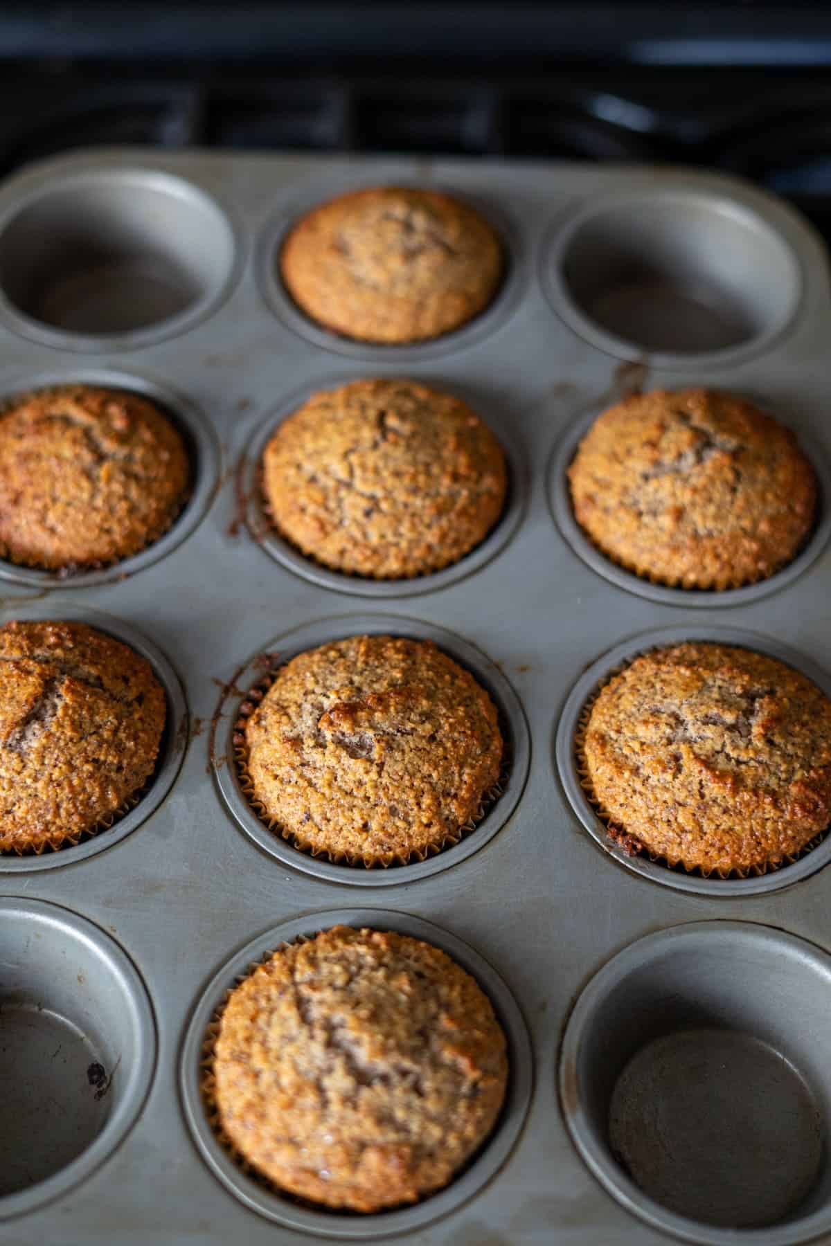 A muffin tin with five plain muffins, two delicious hazelnut muffins, and two empty slots sits atop the kitchen stovetop.