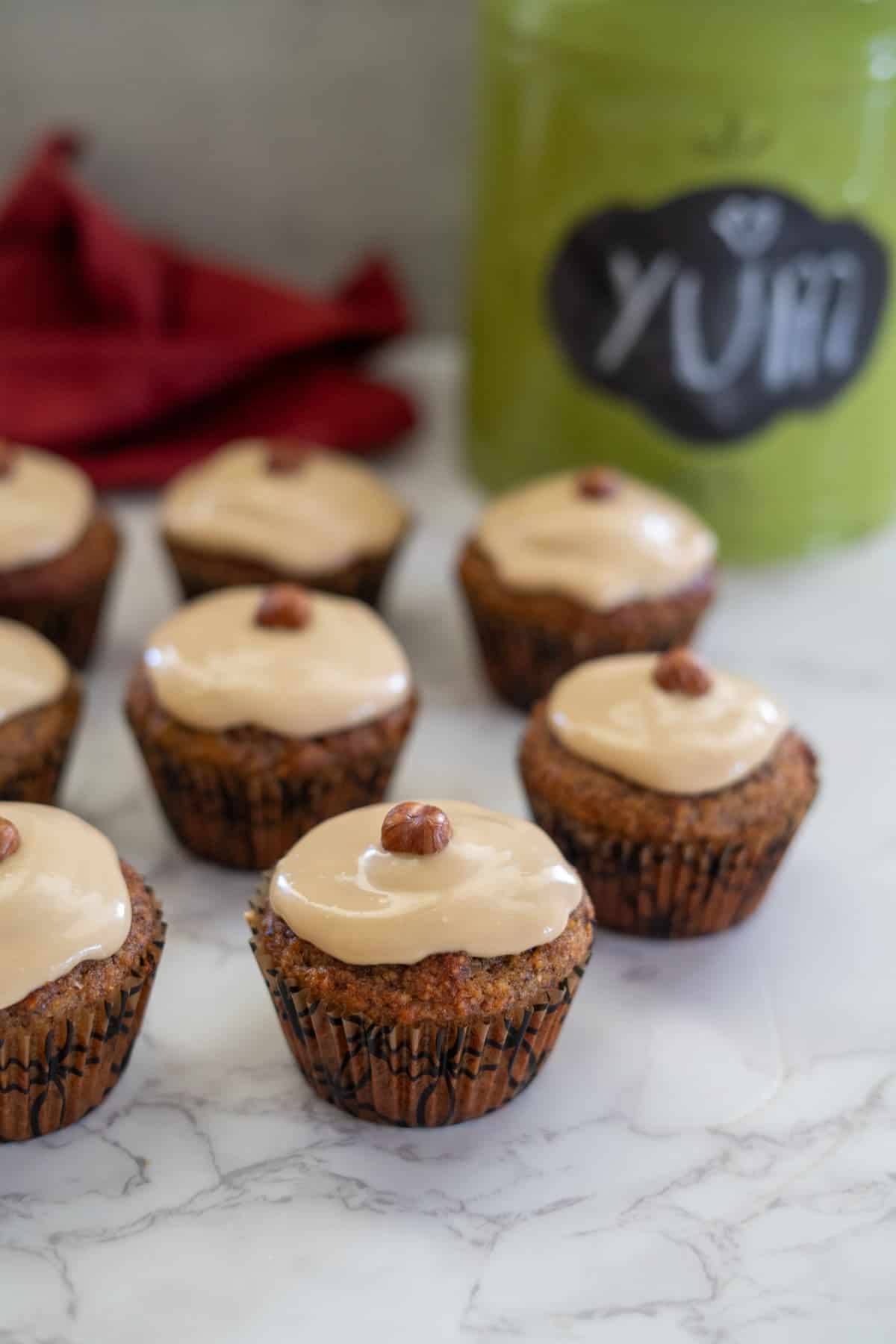 Hazelnut muffins adorned with tan icing and a small nut on top, arranged beautifully on a marble surface. In the background, a green jar labeled "yum" adds an extra touch of charm to this delightful scene.