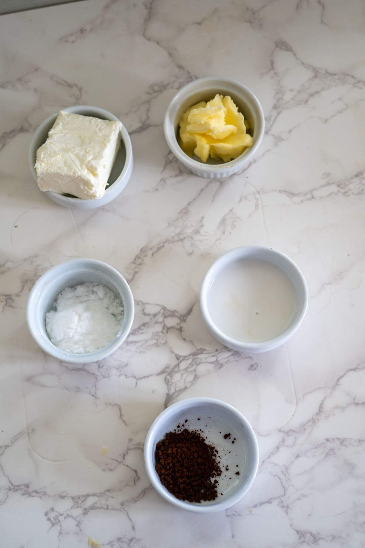 Five small bowls on a marble surface containing cream cheese, butter, cream, powdered sugar, and cocoa powder—perfect companions for freshly baked hazelnut flour muffins.