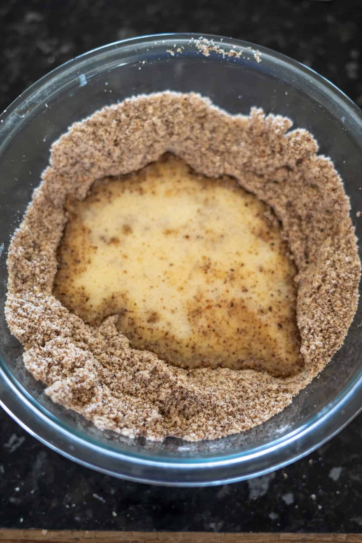 A clear glass bowl with a mixture perfect for hazelnut muffins, featuring brown sugar or flour dusted around the edges and dampened by a creamy liquid pooling in the center.