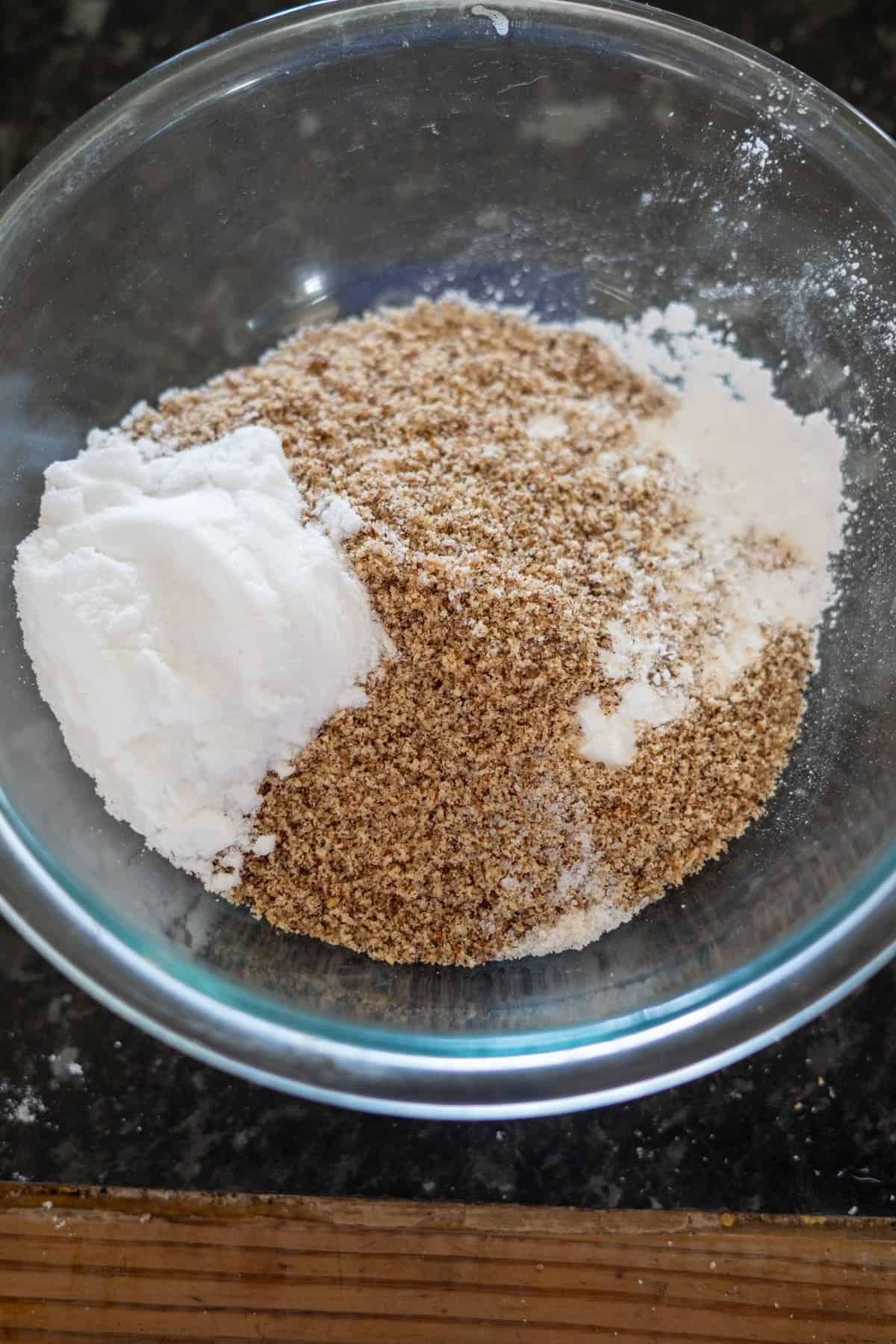 A glass bowl containing almond flour, baking powder, and salt rests on a dark countertop, ready to create delicious hazelnut muffins.