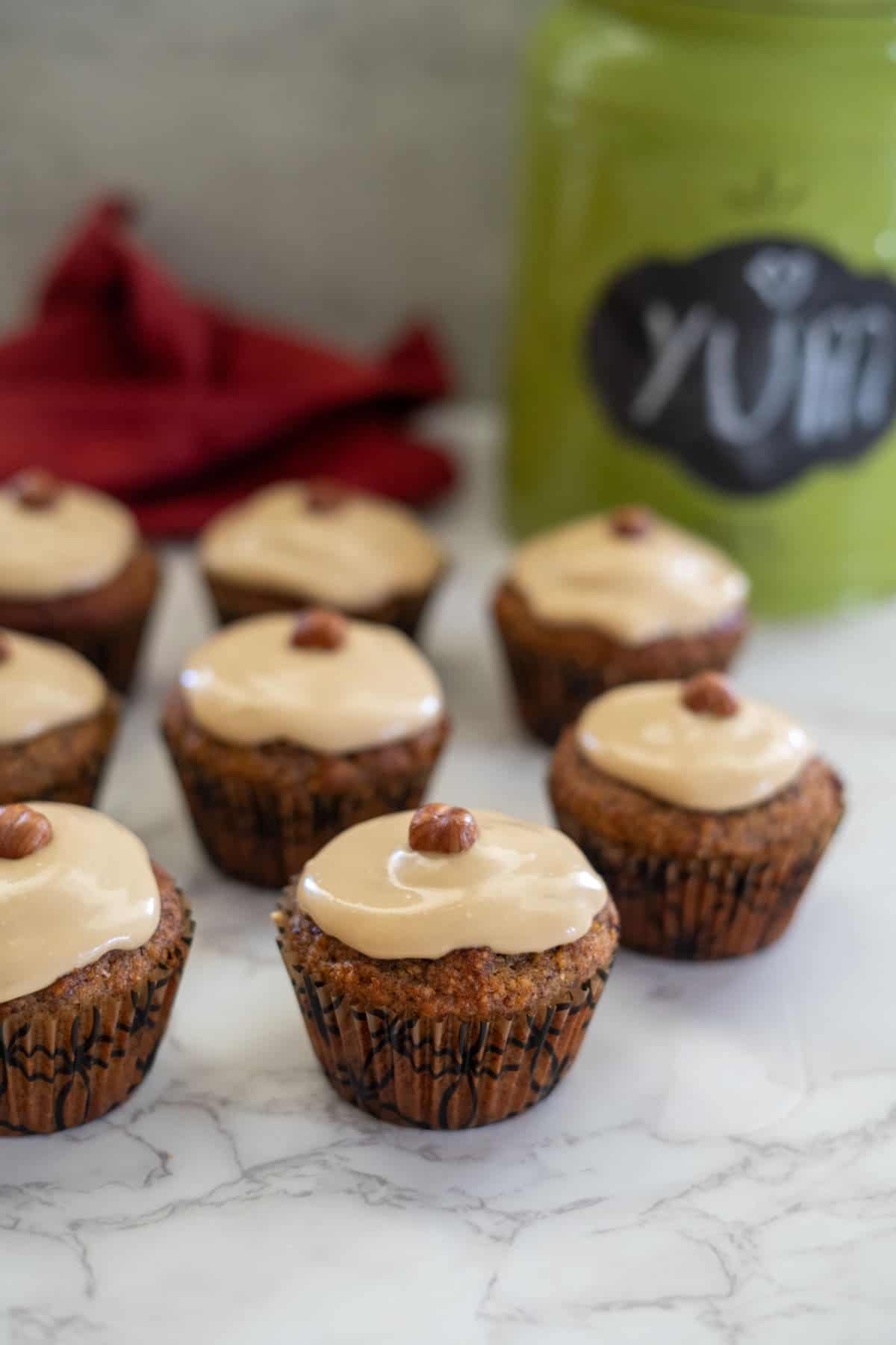 Seven hazelnut muffins, each crowned with a glaze and a single nut, are arranged on a marble surface. A green container bearing the word "YUM" and a red cloth add vibrant touches to the background.