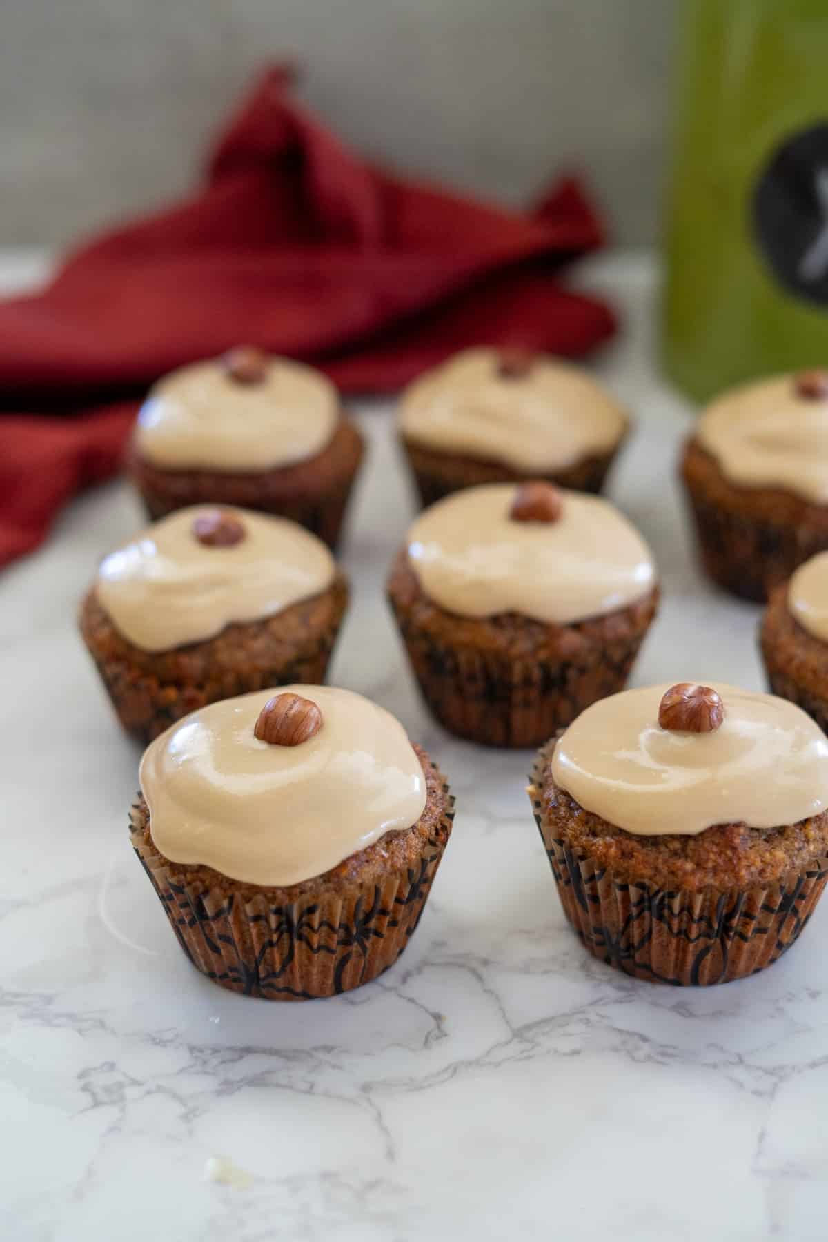 Several cupcakes, crafted with hazelnut flour and topped with light brown frosting and a whole hazelnut on each, are arranged on a marble surface with a red cloth in the background.