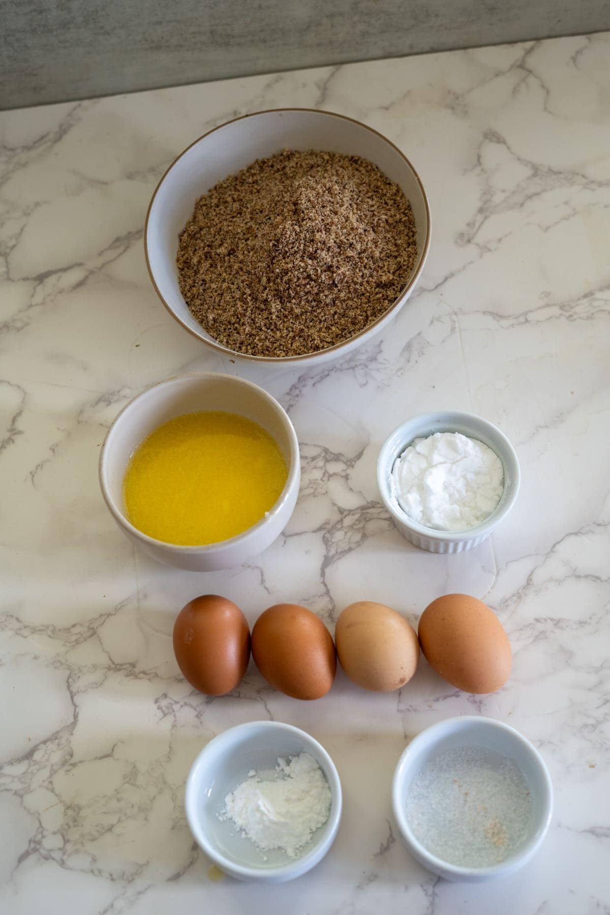 Ingredients arranged on a marble countertop include a bowl of ground almonds, melted butter, eggs, a small bowl of baking soda, another bowl of salt, and the perfect touch for hazelnut flour muffins.