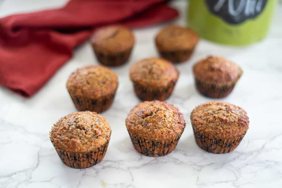 Eight freshly baked hazelnut muffins are artfully arranged on a marble surface, with a vibrant red cloth and a green container adding pops of color in the background.