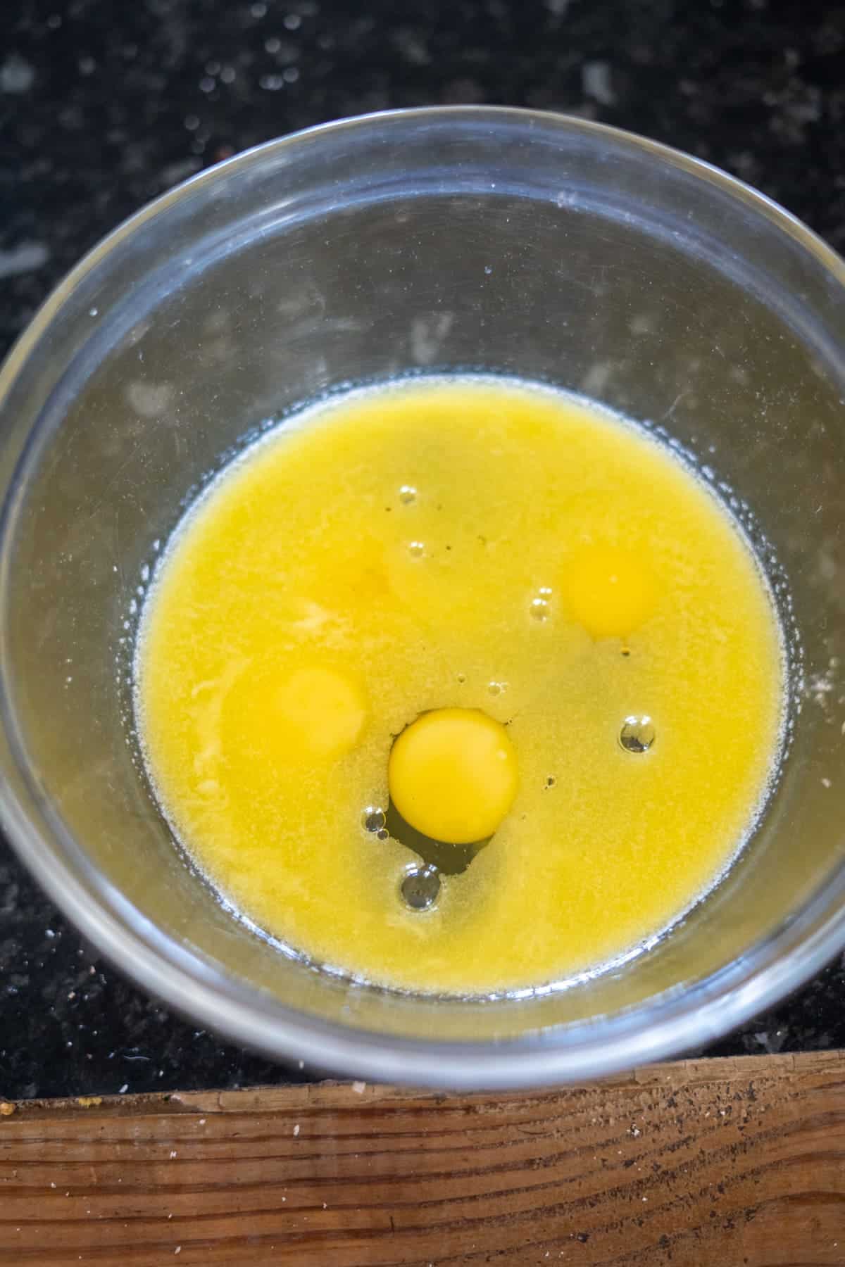 A clear glass bowl contains three cracked eggs on a dark countertop, ready to be transformed into delicious hazelnut muffins.