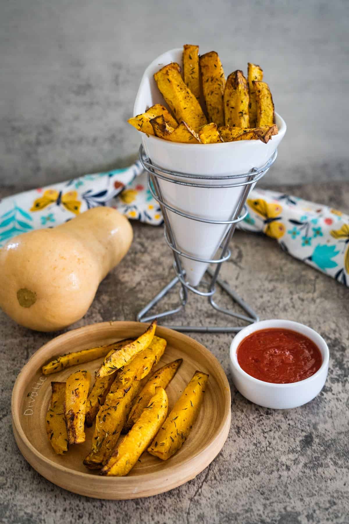 A serving of seasoned butternut squash fries in a white cone holder, with additional fries on a wooden plate next to a small bowl of red dipping sauce, and a butternut squash in the background.