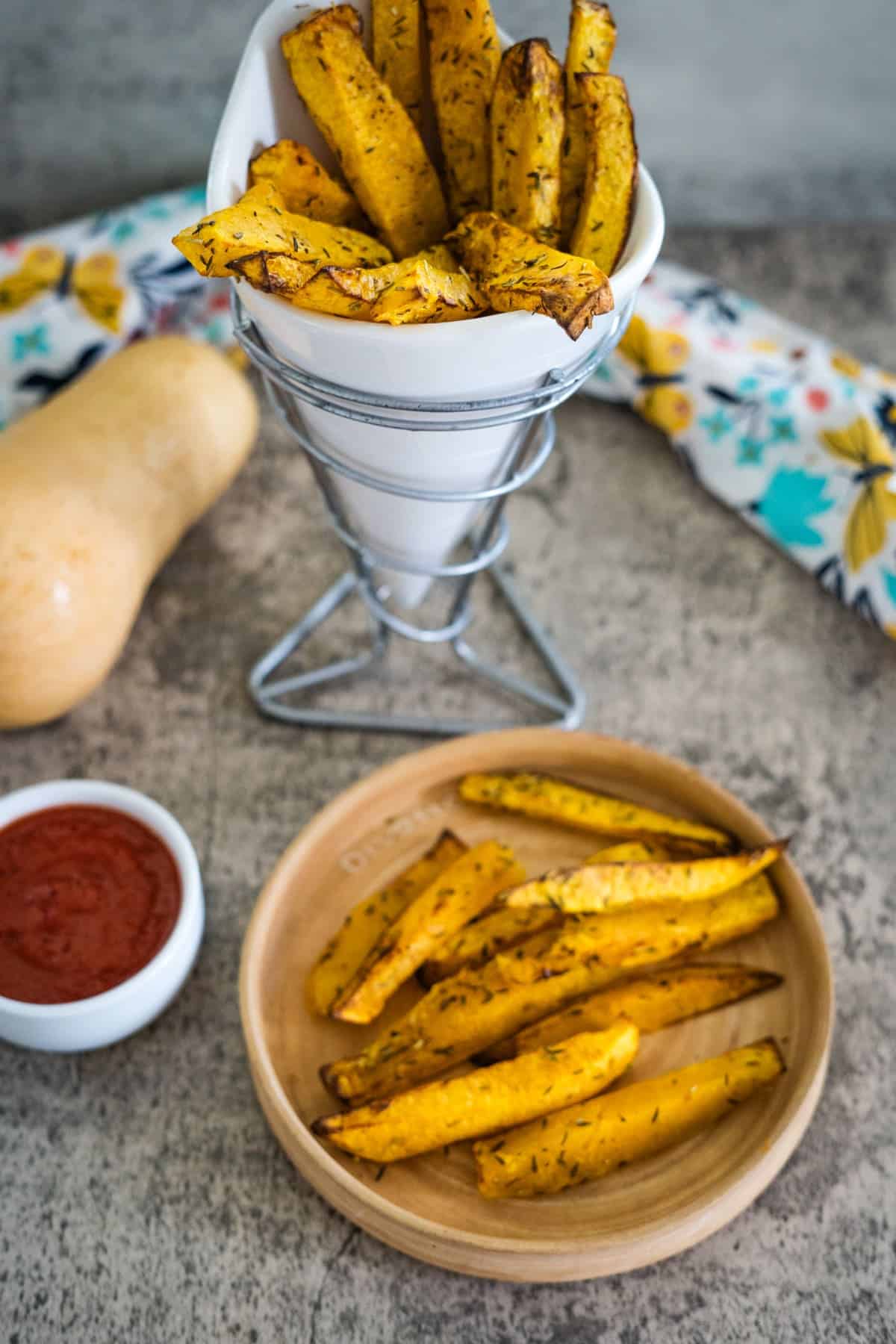 A plate and a cone holder with crispy butternut squash fries, accompanied by a small bowl of red dipping sauce, rest on a textured surface. A raw butternut squash is in the background.