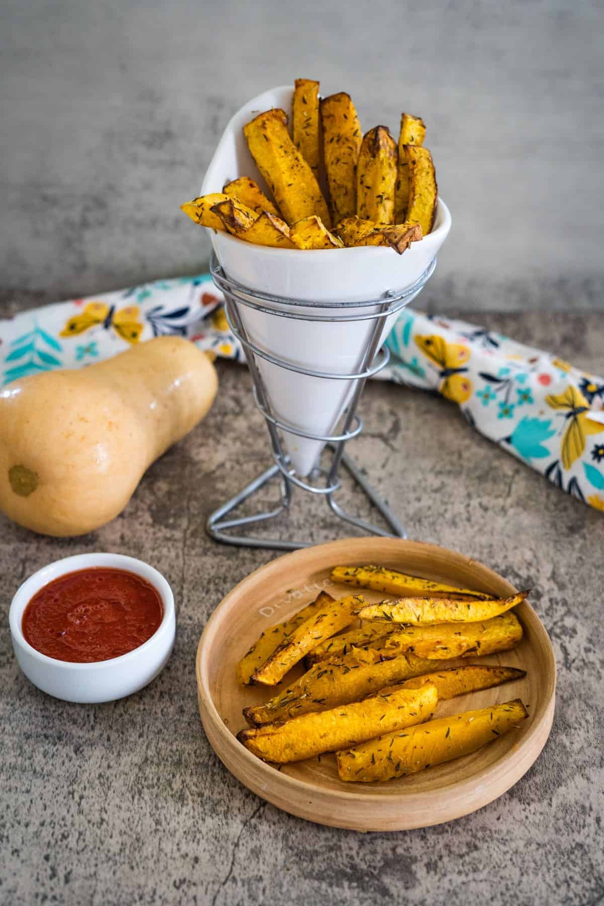 A cone holds butternut squash fries, with a small dish of red dipping sauce and a wooden plate containing more butternut squash fries in the foreground. A whole butternut squash and patterned cloth are in the background.