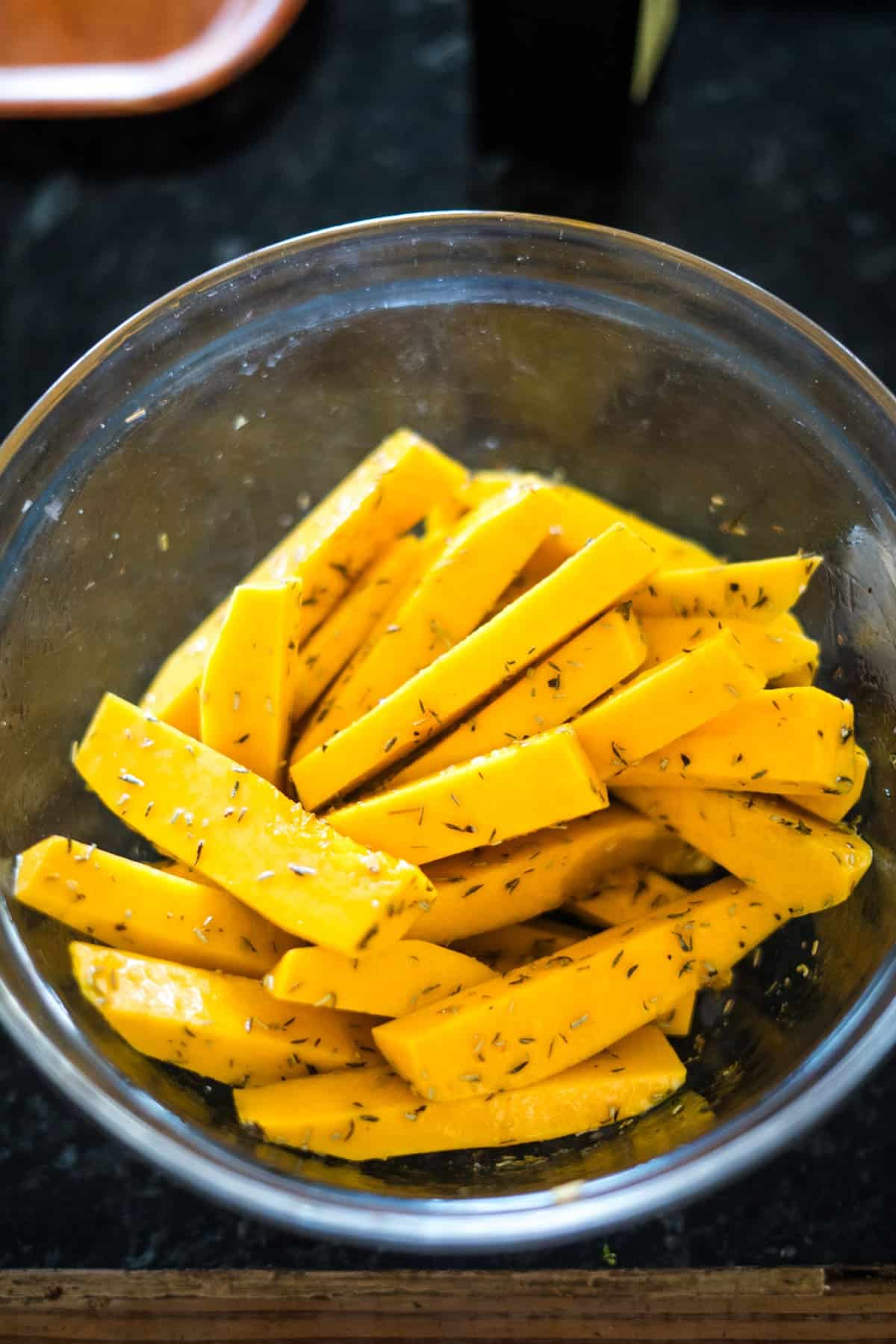 A clear glass bowl filled with raw, seasoned butternut squash fries rests on a dark countertop.