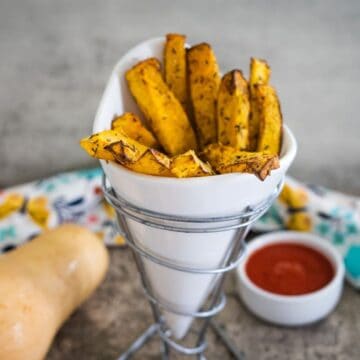 A white cone-shaped container holds butternut squash fries seasoned to perfection. A small bowl of tantalizing dipping sauce awaits in the background.