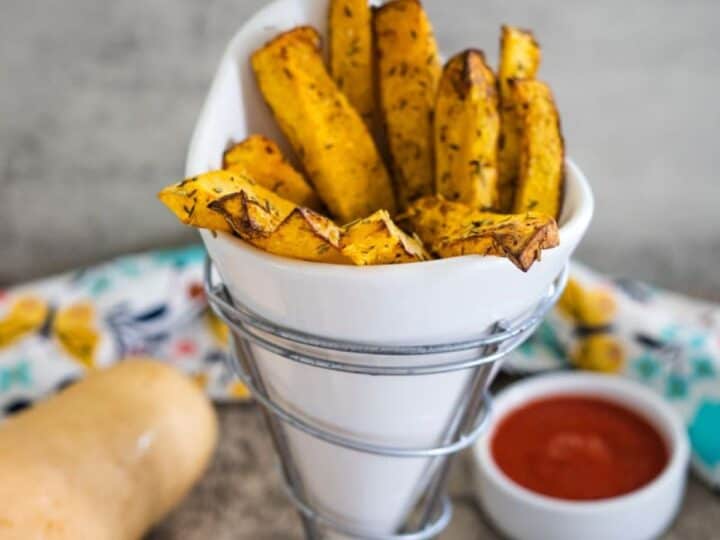 A white cone-shaped container holds butternut squash fries seasoned to perfection. A small bowl of tantalizing dipping sauce awaits in the background.