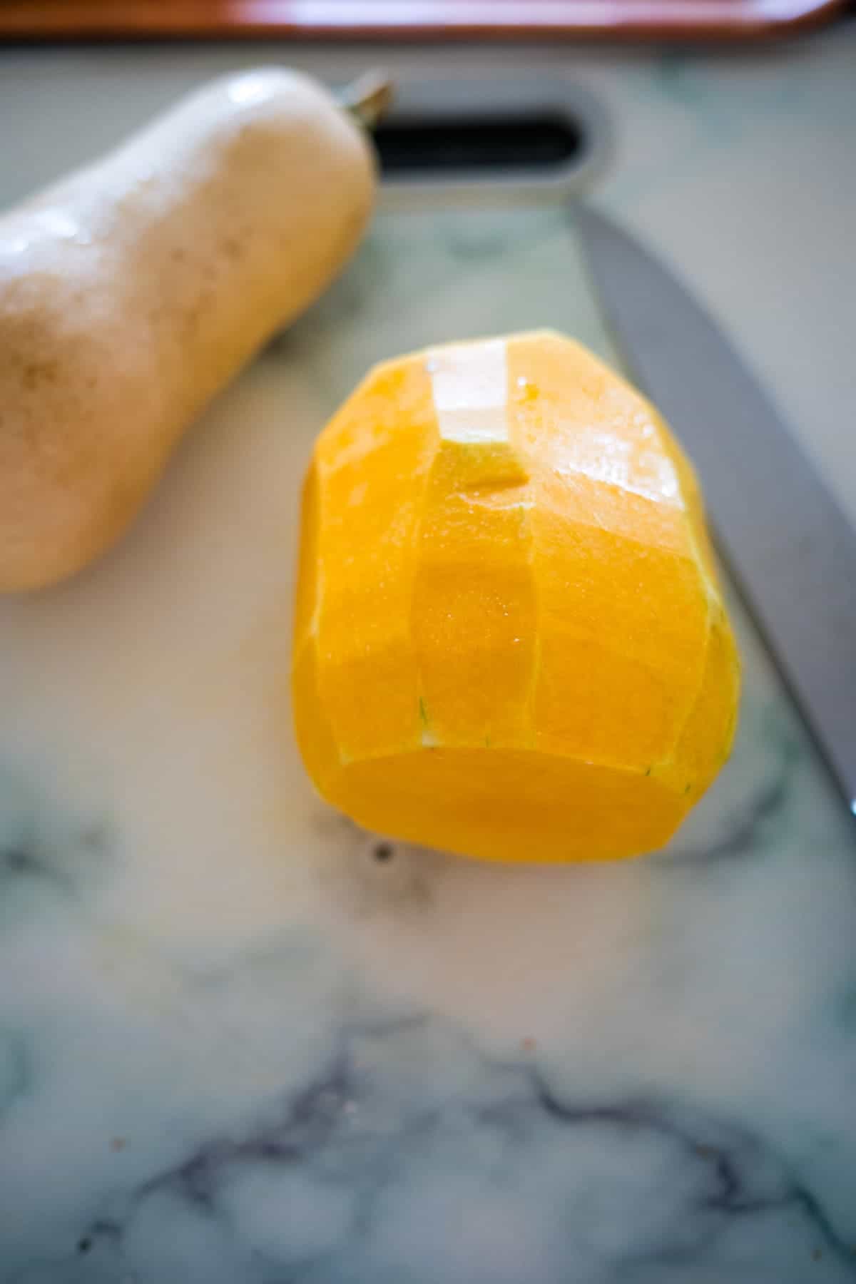 A partially peeled butternut squash, perfect for making butternut squash fries, is placed beside a knife on a marble countertop.