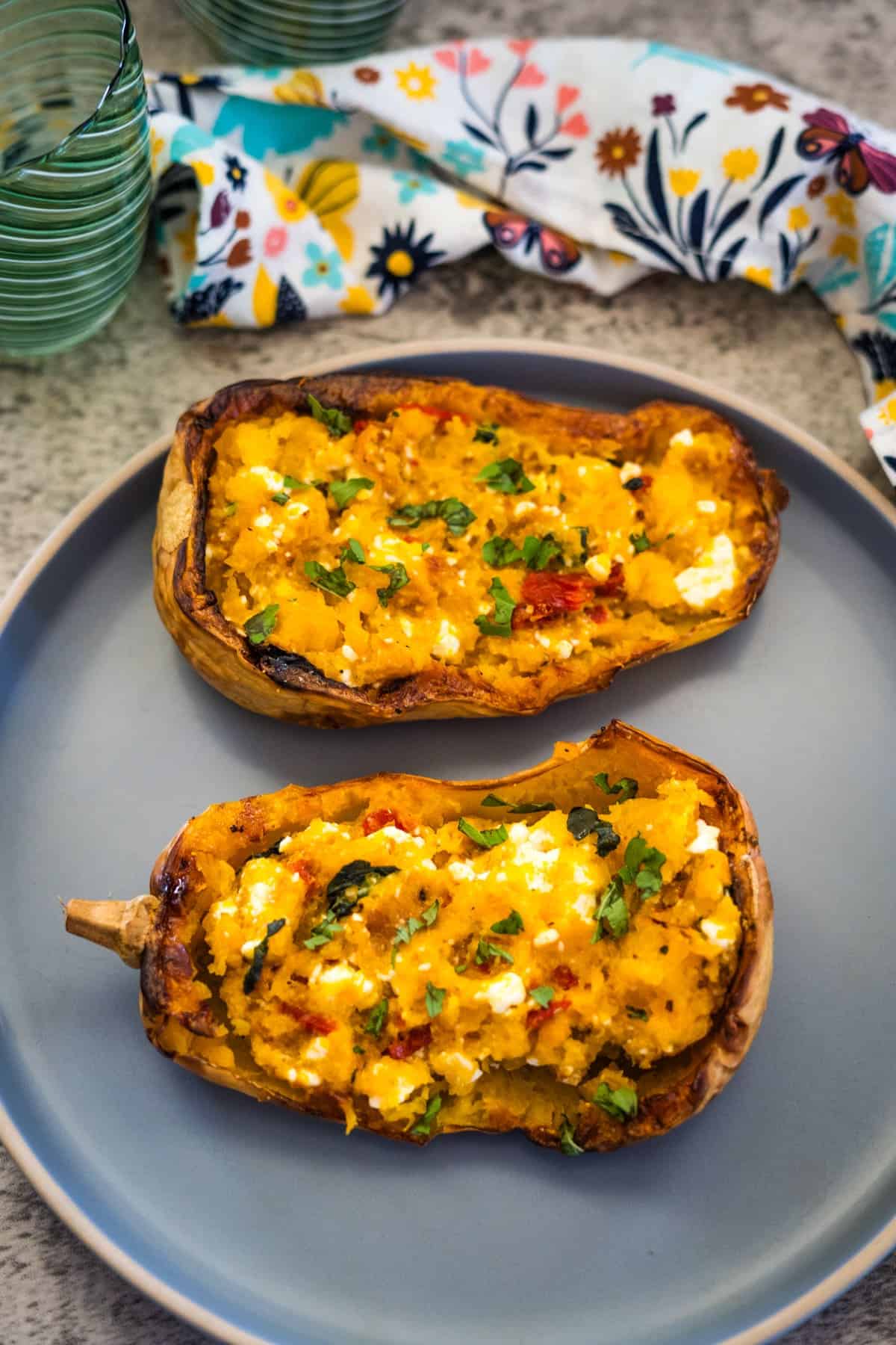 Two baked butternut squash halves filled with a seasoned mixture and crumbled feta, garnished with herbs, served on a gray plate, with a floral cloth and two green glasses in the background.