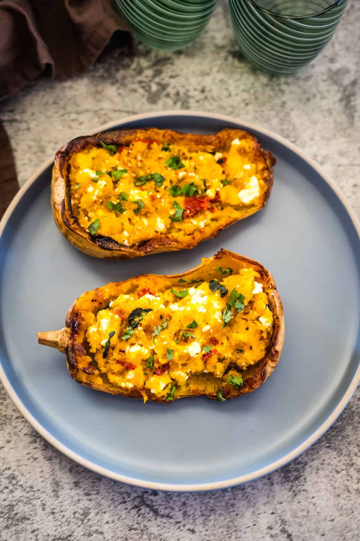 Two halves of a Feta Stuffed Butternut Squash with mixed ingredients, garnished with herbs, placed on a grey plate on a grey surface. Two empty green glasses are visible in the background.