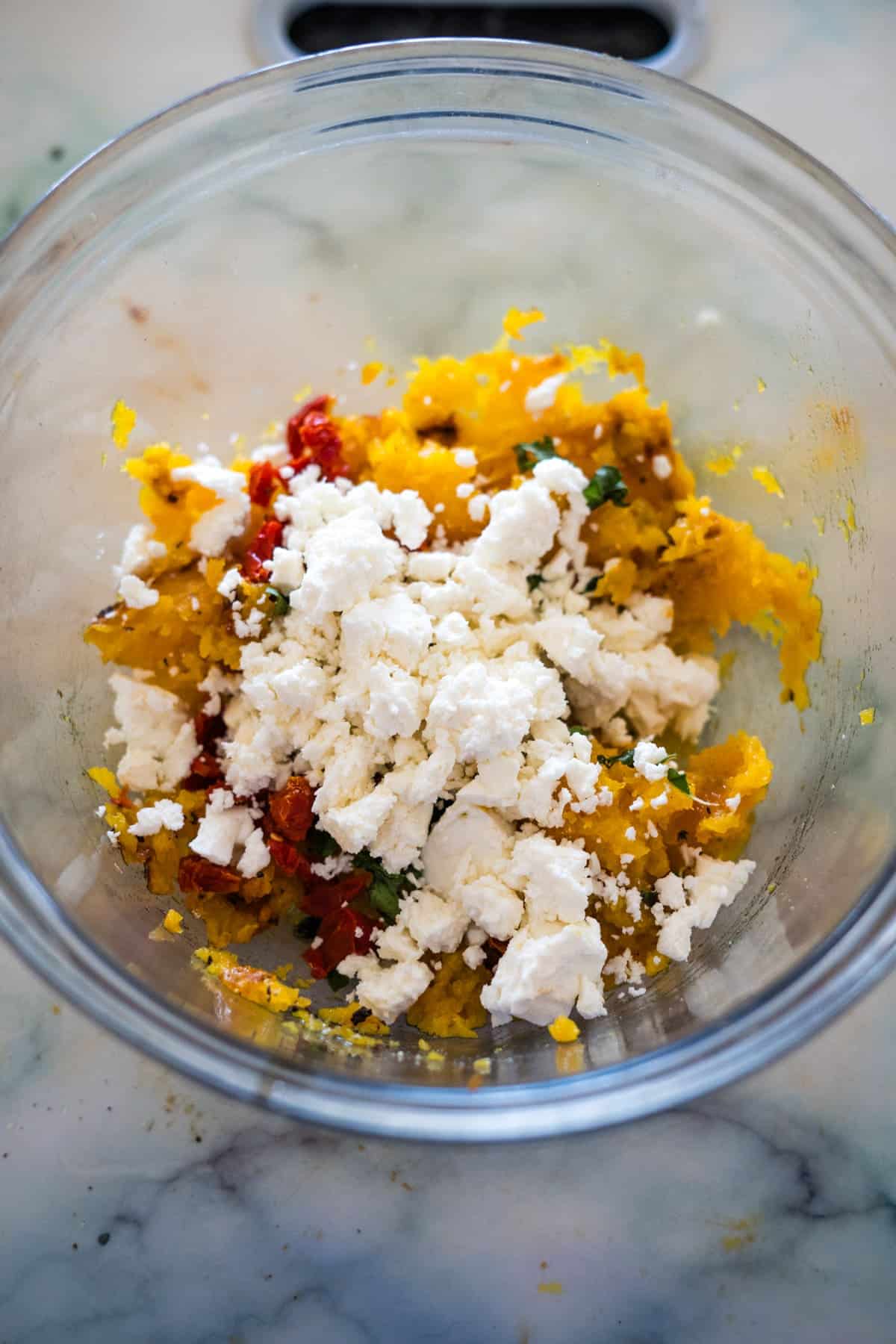 A glass bowl containing a delightful mixture of mashed butternut squash, crumbled feta cheese, and chopped red bell peppers, perfect for creating appetizing Feta Stuffed Butternut Squash.