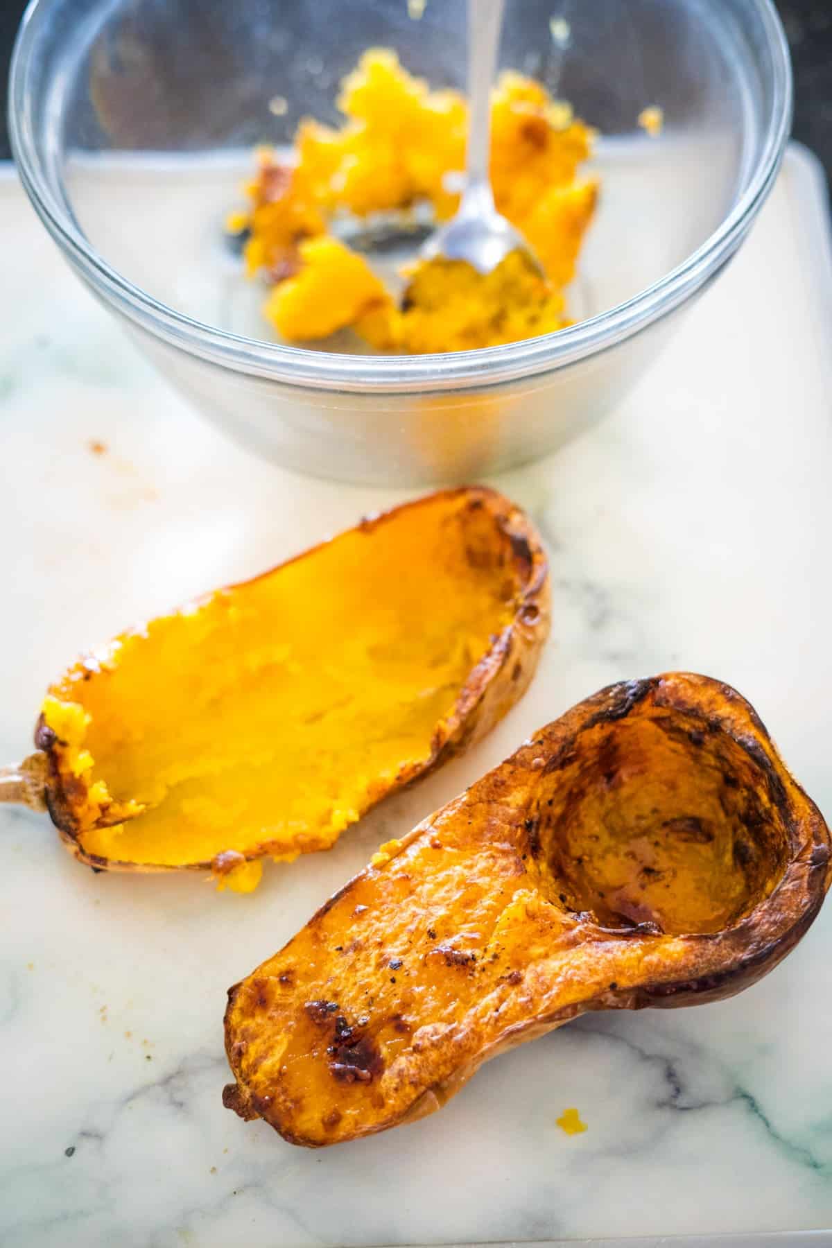 Two roasted butternut squash halves lie on a white surface, with one half partially scooped out. A bowl containing mashed squash and a spoon is in the background, hinting at the delicious preparation of feta stuffed butternut squash.