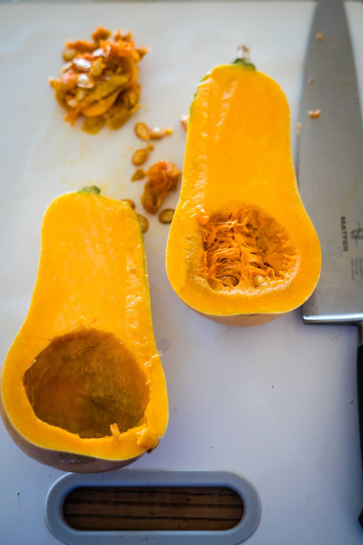 A butternut squash cut in half, with seeds removed and placed next to a knife on a white cutting board, ready to become delicious Feta Stuffed Butternut Squash.