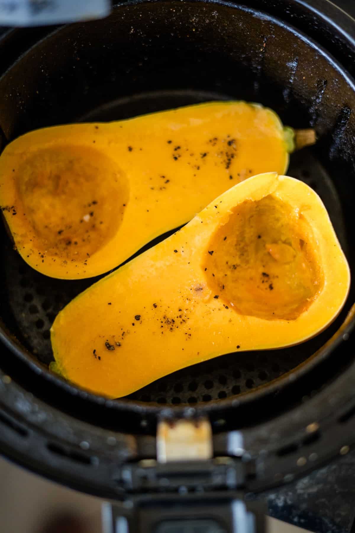 Two halves of a butternut squash, seasoned with salt and pepper, are placed inside an air fryer basket, ready for cooking. Imagine the delicious aroma as they roast to perfection, creating the ideal base for Feta Stuffed Butternut Squash.