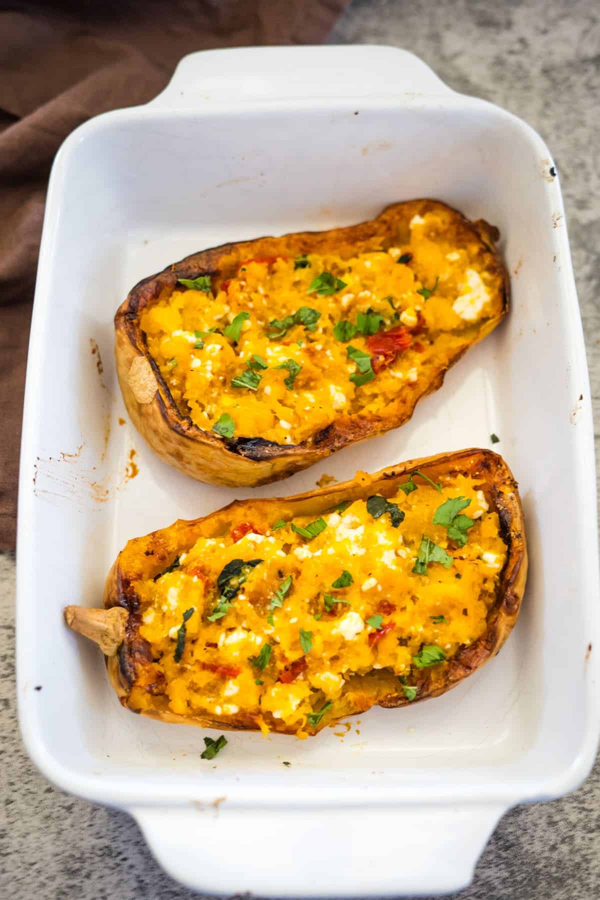 A white baking dish containing two halves of Feta Stuffed Butternut Squash, topped with chopped herbs and melted cheese.