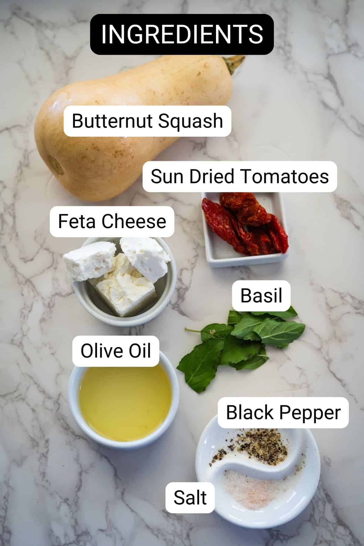 Ingredients for Feta Stuffed Butternut Squash include butternut squash, sun-dried tomatoes, feta cheese, basil, olive oil, black pepper, and salt—all arranged on a marble surface.