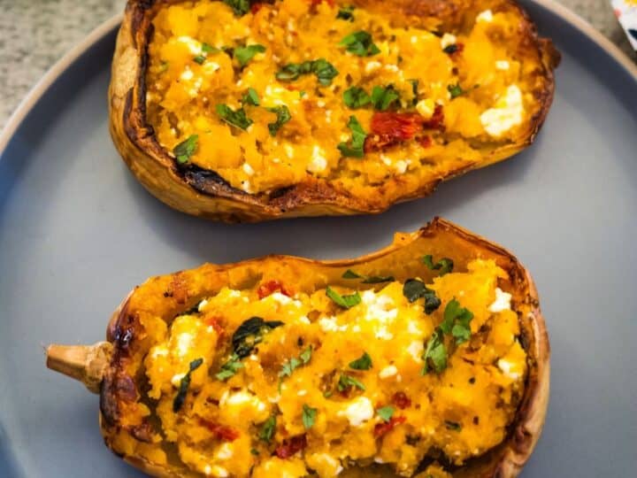 Two halves of a baked stuffed butternut squash filled with a mixture of mashed squash, herbs, and cheese are served on a gray plate.