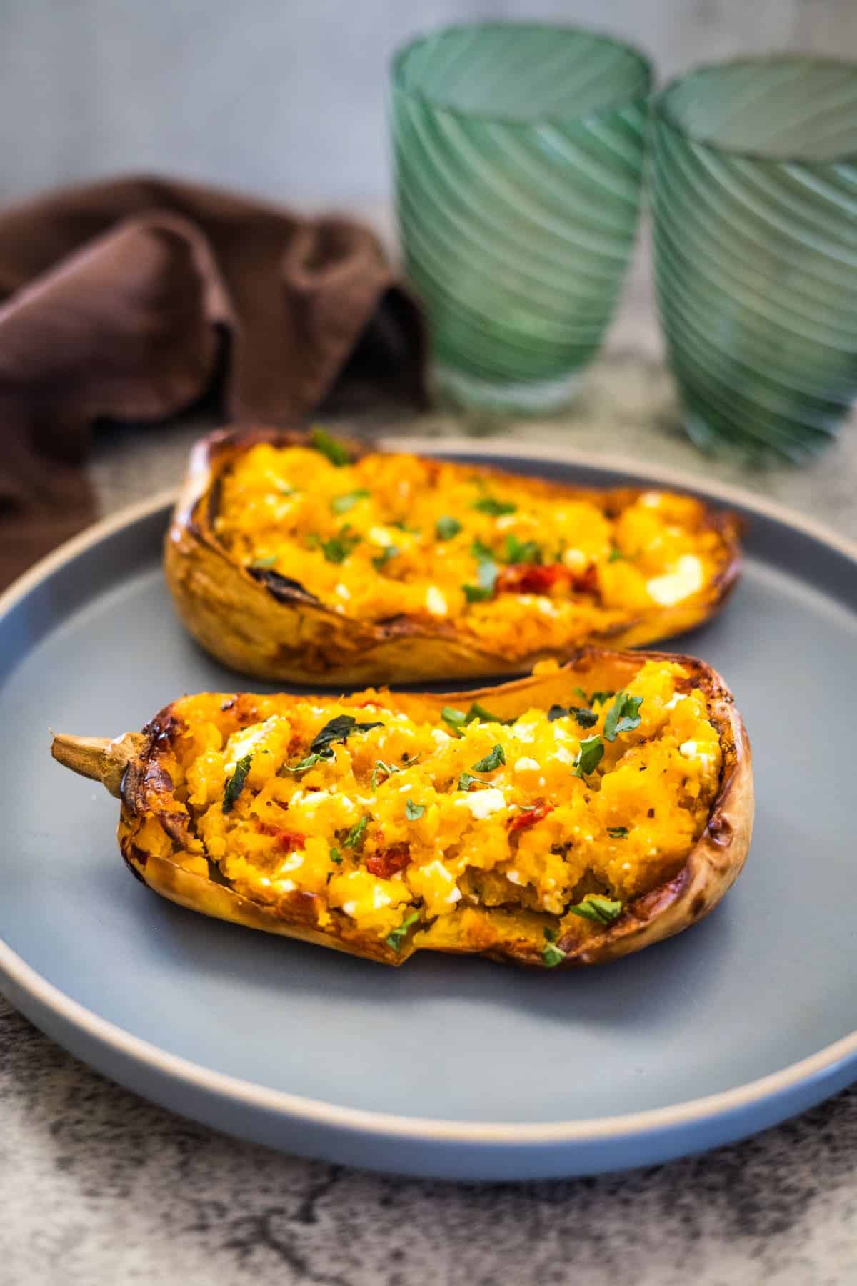 Two stuffed eggplant halves topped with herbs are served on a blue plate. Two empty green glasses and a brown napkin are in the background, reminiscent of a vibrant Feta Stuffed Butternut Squash presentation.