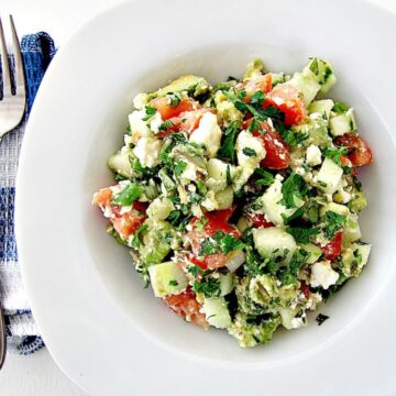 A bowl of avocado salad containing tomatoes, cucumbers, feta cheese, and herbs, presented on a white background.
