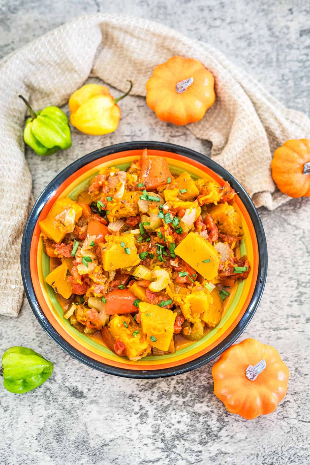 A bowl of cooked mixed vegetables sits on a textured surface, garnished with chopped herbs. Small ornamental pumpkins and peppers are placed around the bowl, including a vibrant Caribbean Spiced Pumpkin. A beige cloth is draped in the background.