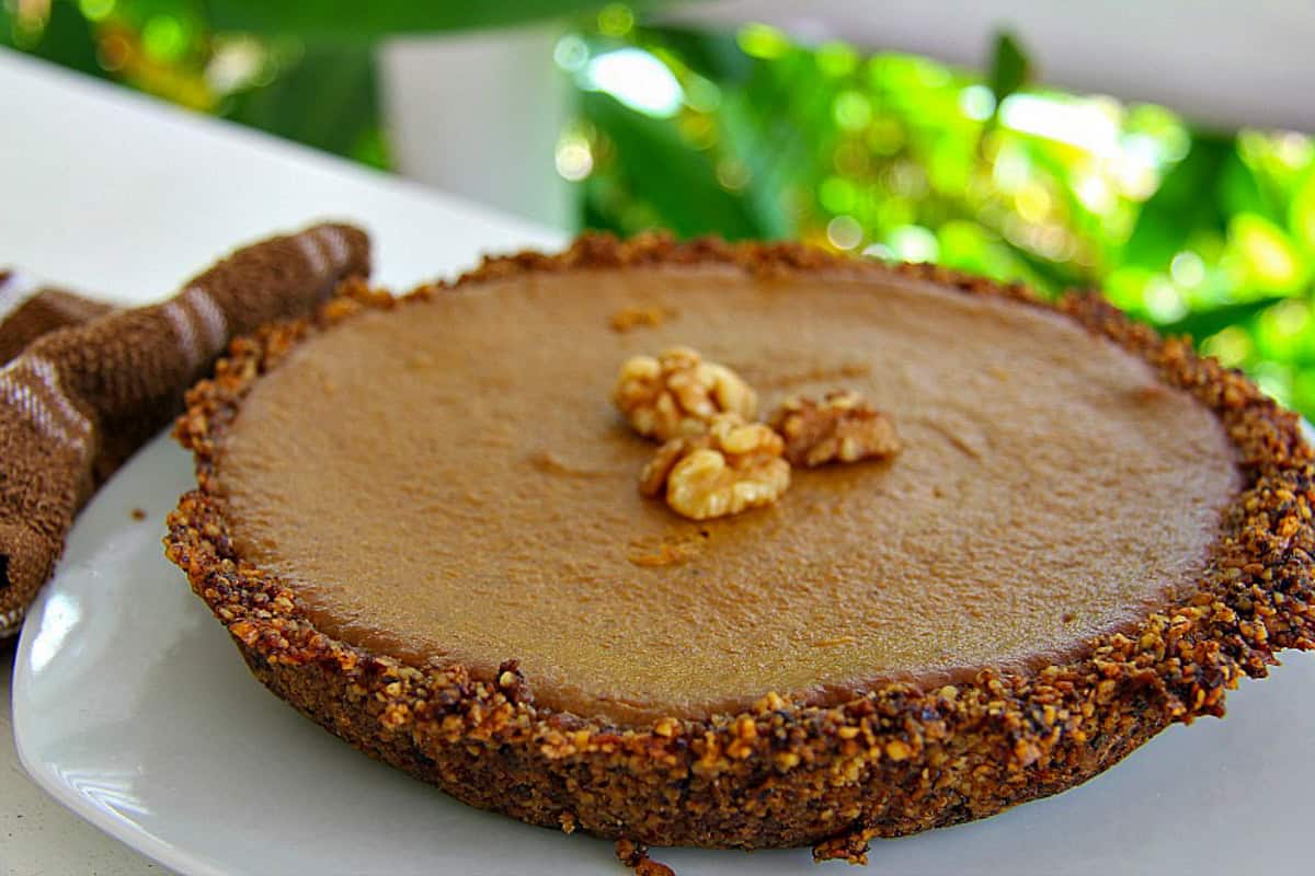 A round walnut pumpkin pie with a nut-filled crust and smooth filling, topped generously with walnuts, sits elegantly on a white plate. Soft green leaves fade into the background, adding a touch of serenity to this delightful scene.