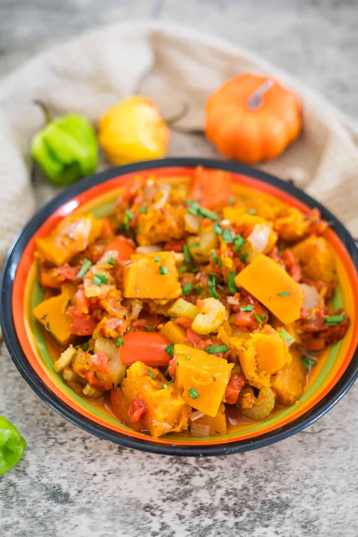 A colorful bowl of Caribbean Spiced Pumpkin stew with vegetables, garnished with herbs, sits on a table next to decorative pumpkins and peppers.