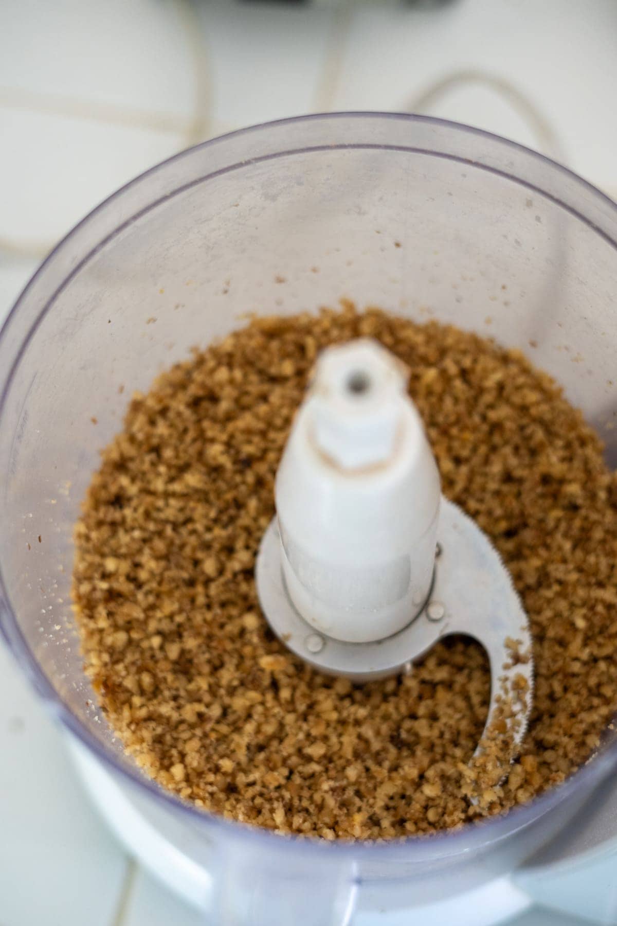 Chopped walnuts inside a food processor bowl with a metal blade attachment visible, ready to infuse your pumpkin pie with a delightful crunch.