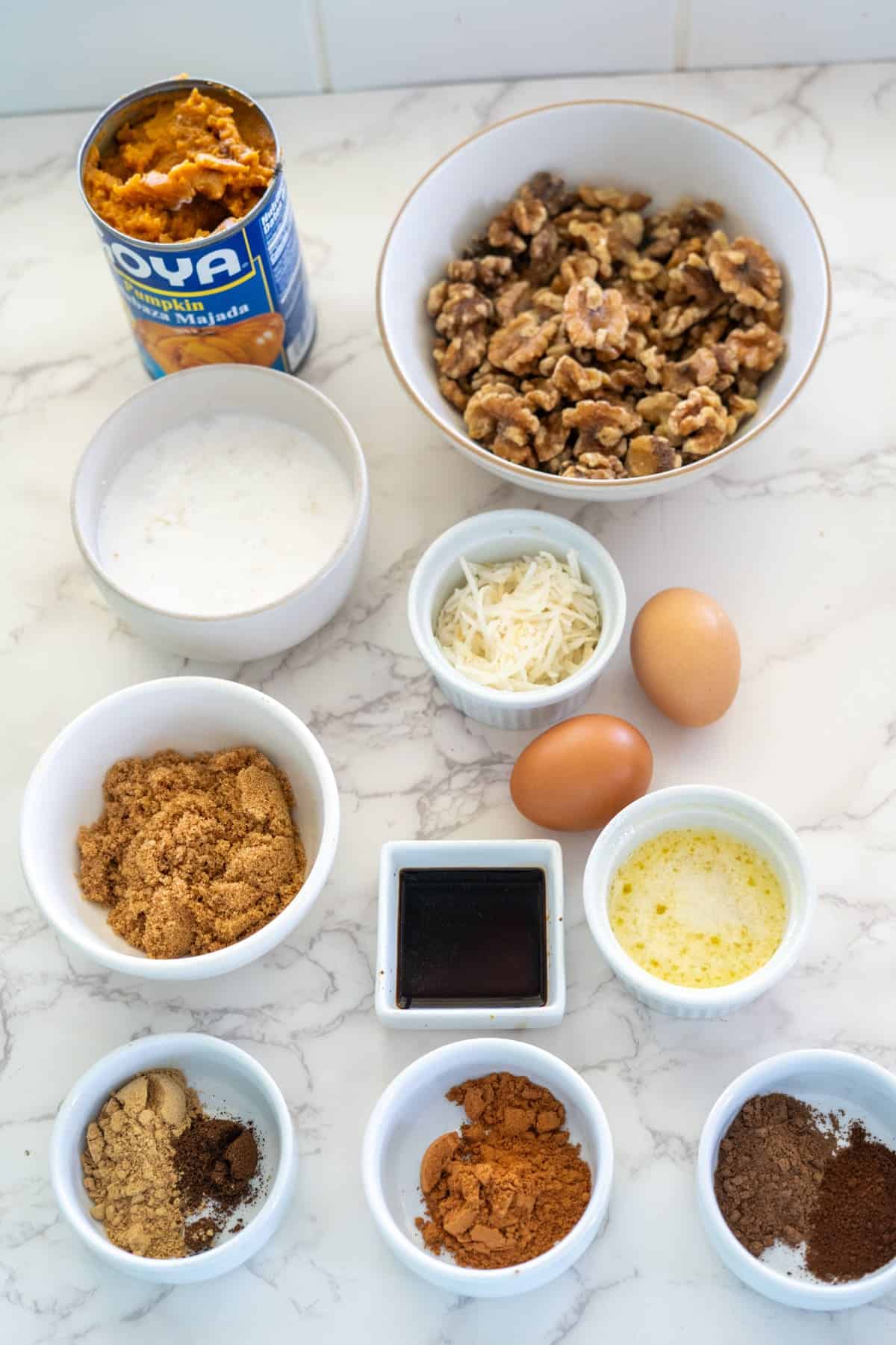 Ingredients for a walnut pumpkin pie are beautifully arranged on a marble surface. They include canned pumpkin, chopped walnuts, milk, eggs, brown sugar, shredded coconut, vanilla extract, butter, and an assortment of spices neatly presented in small bowls.