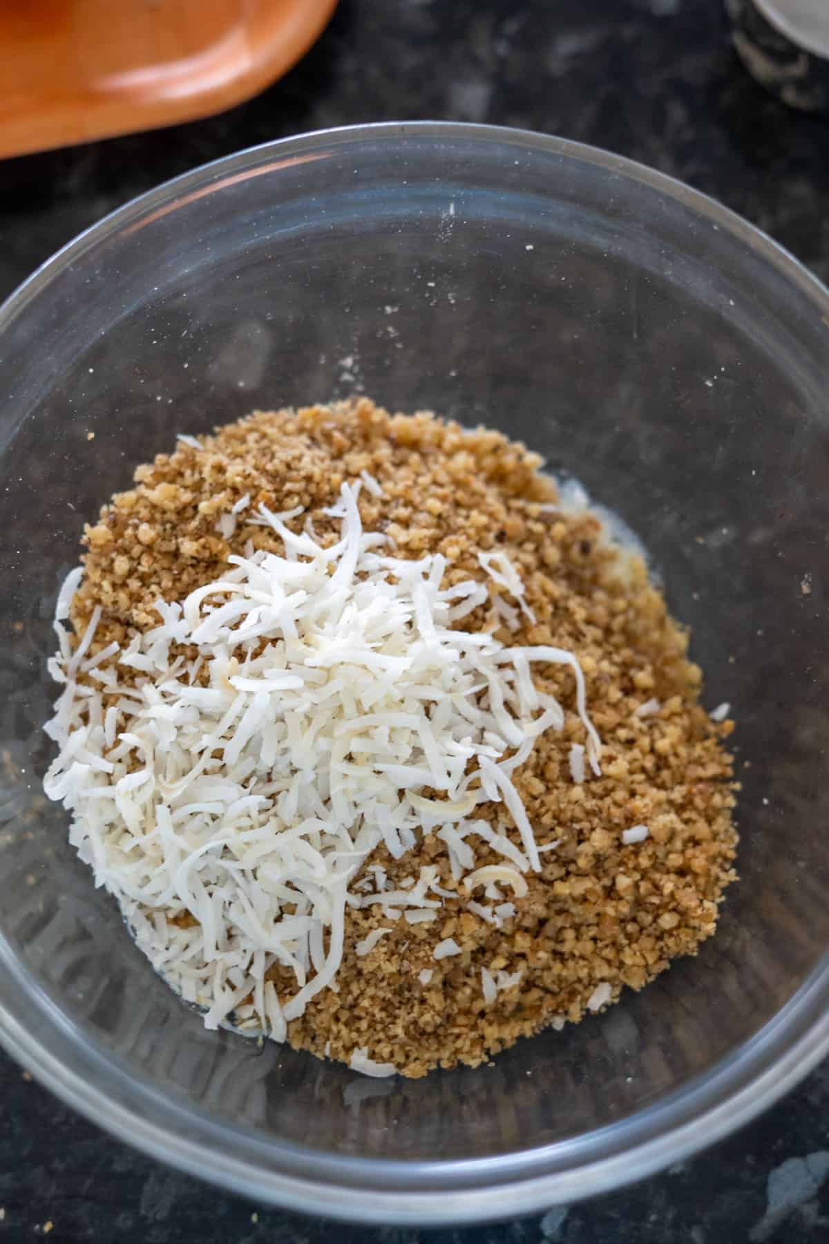 A clear bowl containing shredded coconut and crushed graham crackers rests invitingly on a black countertop, reminiscent of the textures found in a delicious walnut pumpkin pie.