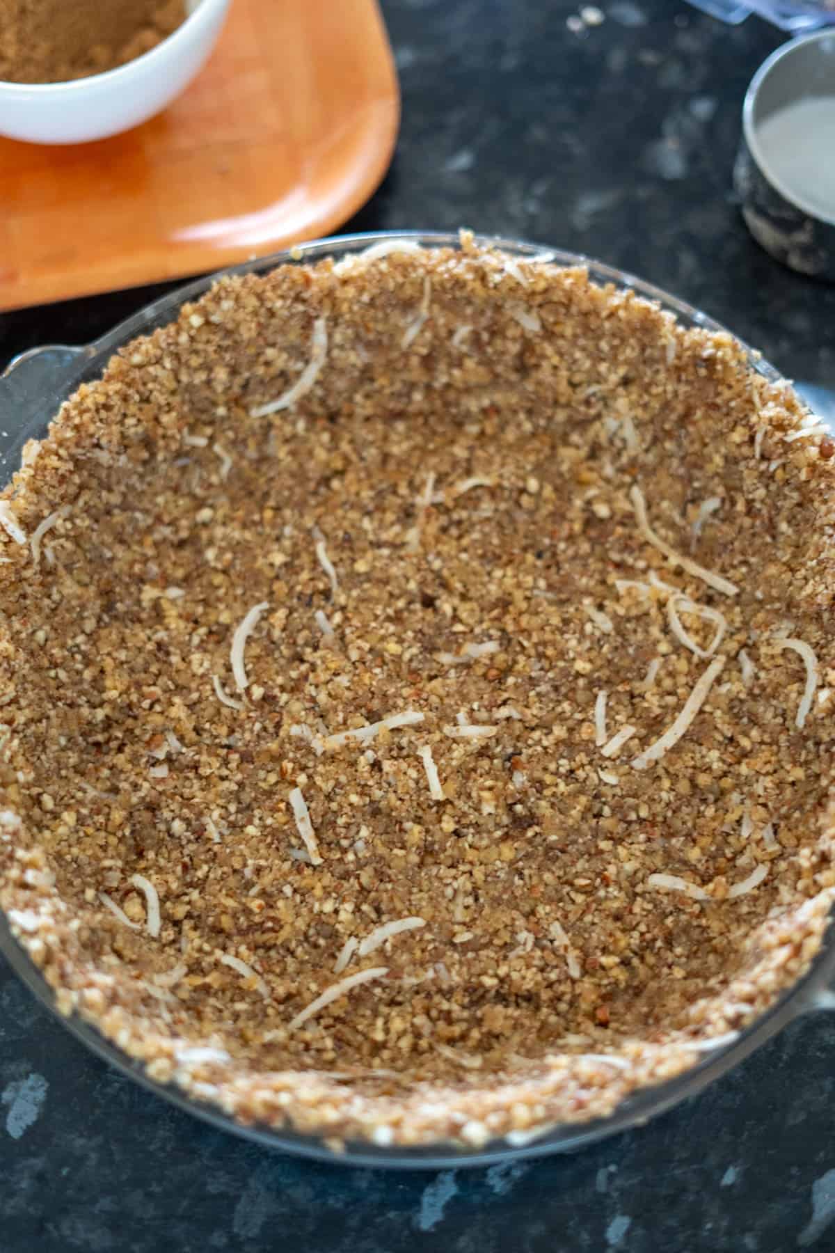 A pie crust filled with a mixture of finely chopped walnuts and shredded coconut is in a glass pie dish on a kitchen counter.