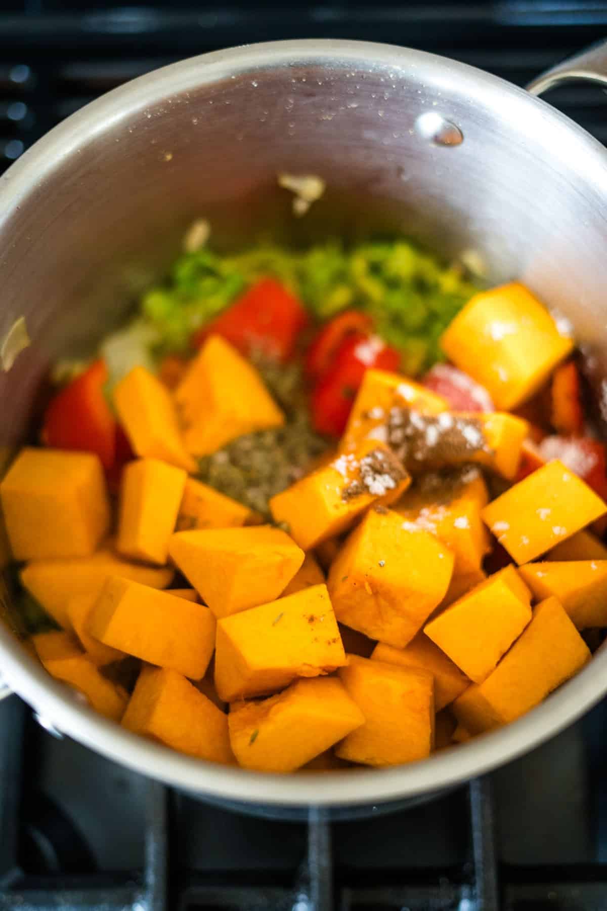 Stainless steel pot on a stove containing chopped butternut squash, bell peppers, Caribbean spiced pumpkin seasonings, and other ingredients being cooked.