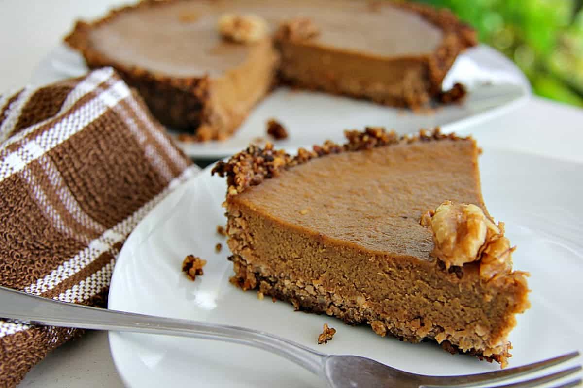 A slice of walnut pumpkin pie on a white plate, crowned with a walnut, sits next to a fork and a brown striped cloth. Another pie awaits in the background.