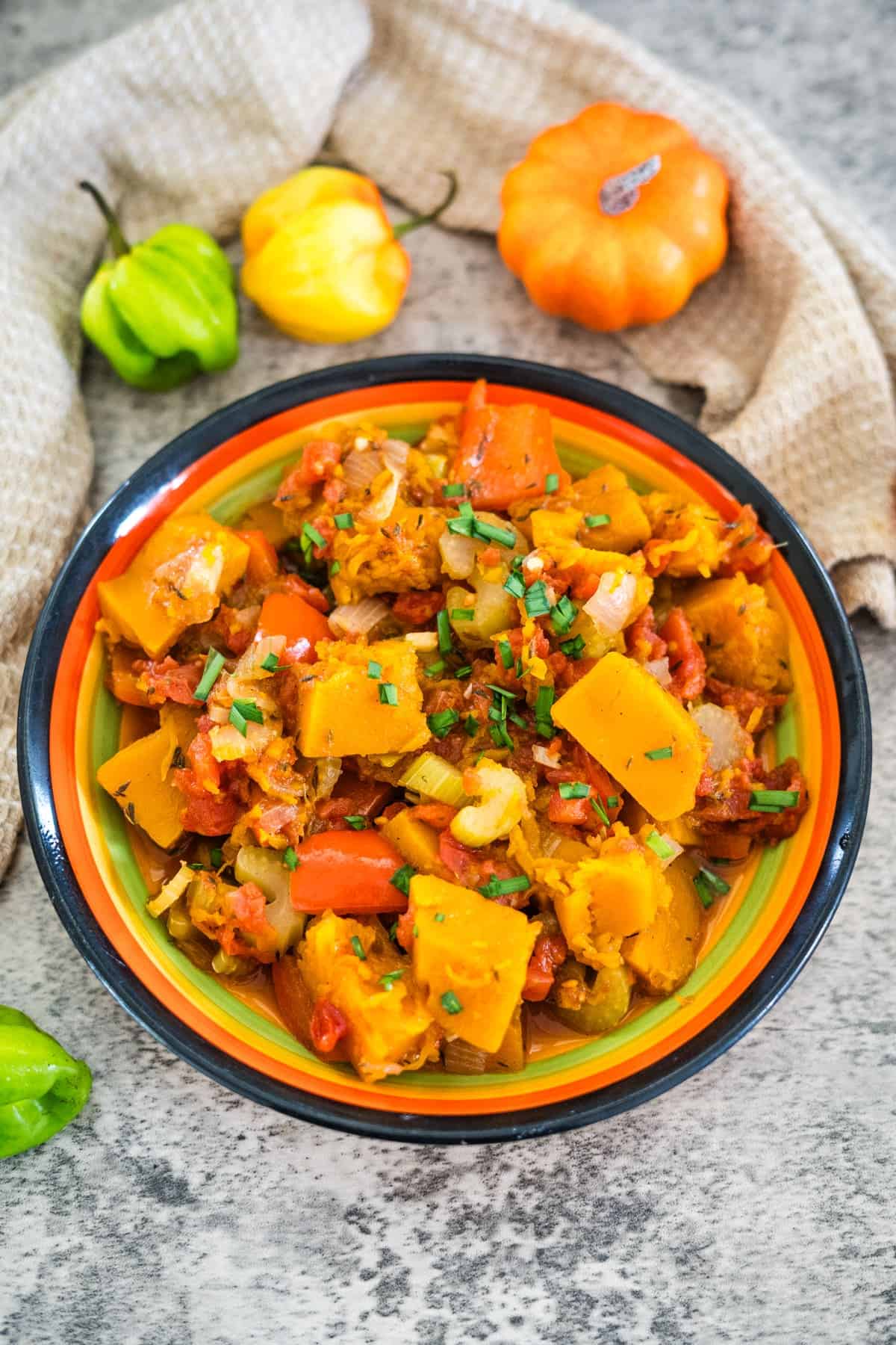 A colorful bowl of cooked vegetables including squash, carrots, tomatoes, and onions, garnished with chopped herbs. Caribbean spiced pumpkin adds a unique twist. Small decorative gourds are in the background.