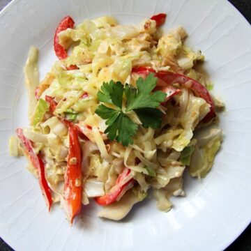 A white plate containing a creamy fried cabbage salad mix with red bell pepper strips, shredded cheese, and a parsley garnish.