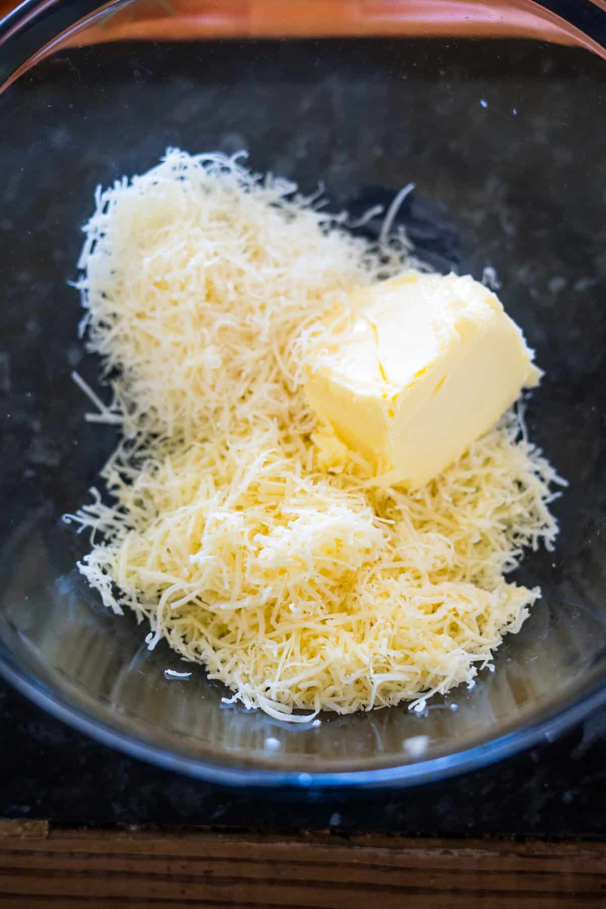 Shredded cheese and a block of butter sit in a glass bowl on a dark countertop, perfect ingredients for making delicious cheese crackers.