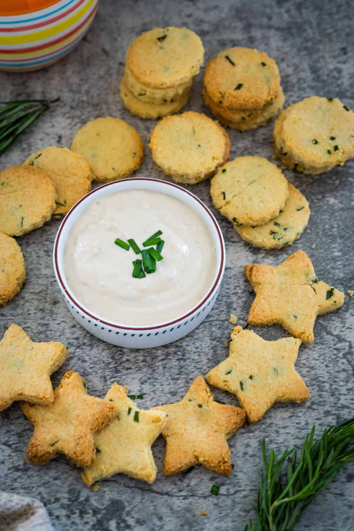 Round and star-shaped herb biscuits and cheese crackers surround a bowl of white dip topped with green garnish on a gray surface.