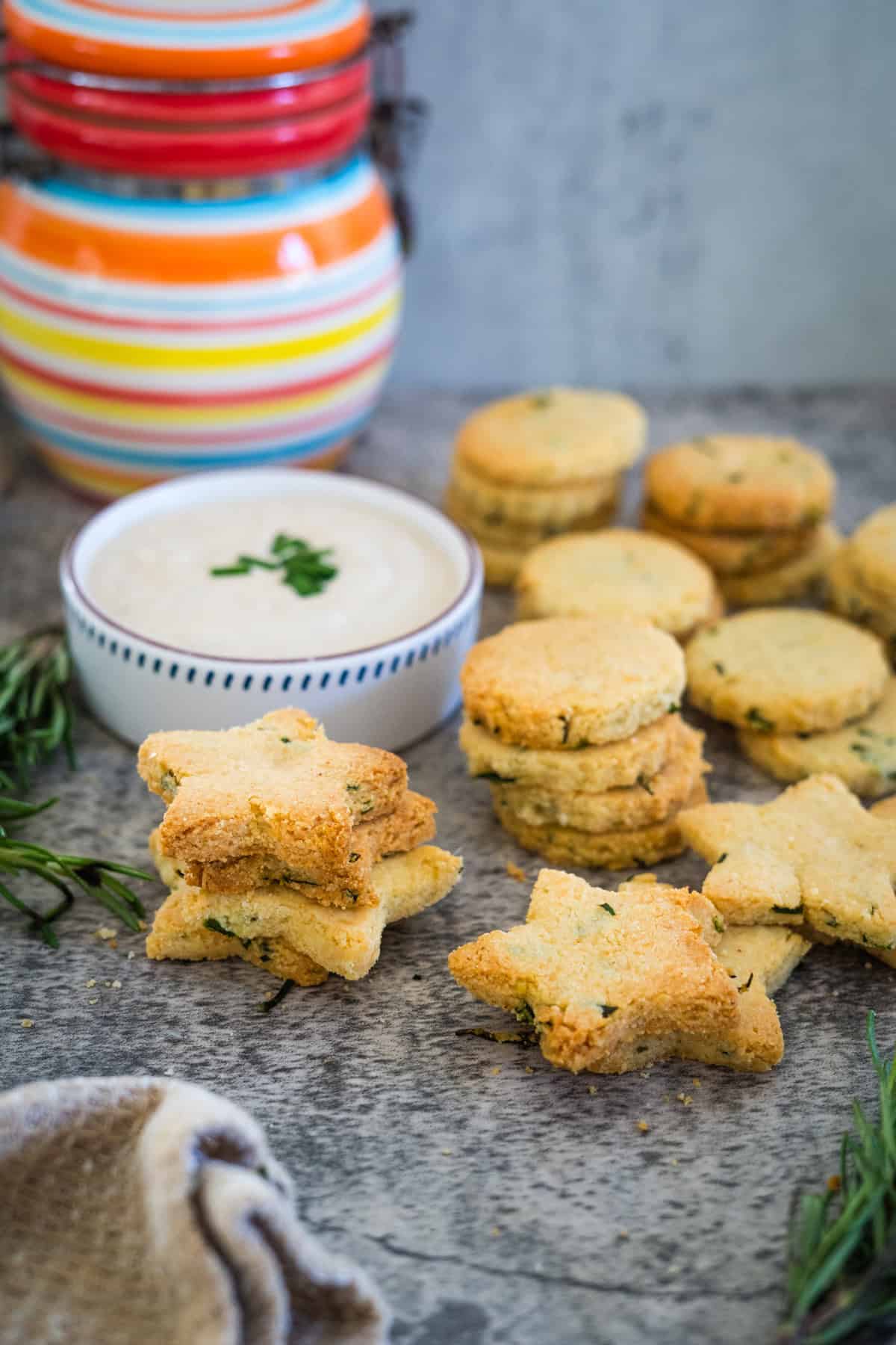 A variety of star-shaped and round cheese crackers sit enticingly beside a bowl of dip in front of a striped jar. Rosemary is artfully scattered around the surface, adding a touch of elegance.