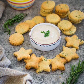 Star and round-shaped cheese crackers with herbs are arranged around a bowl of dip, garnished with chopped chives, on a textured surface.