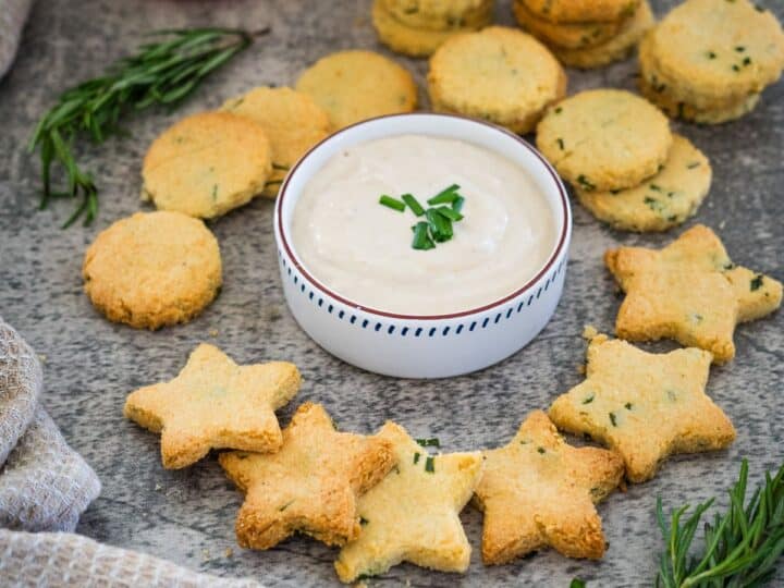 Star and round-shaped cheese crackers with herbs are arranged around a bowl of dip, garnished with chopped chives, on a textured surface.