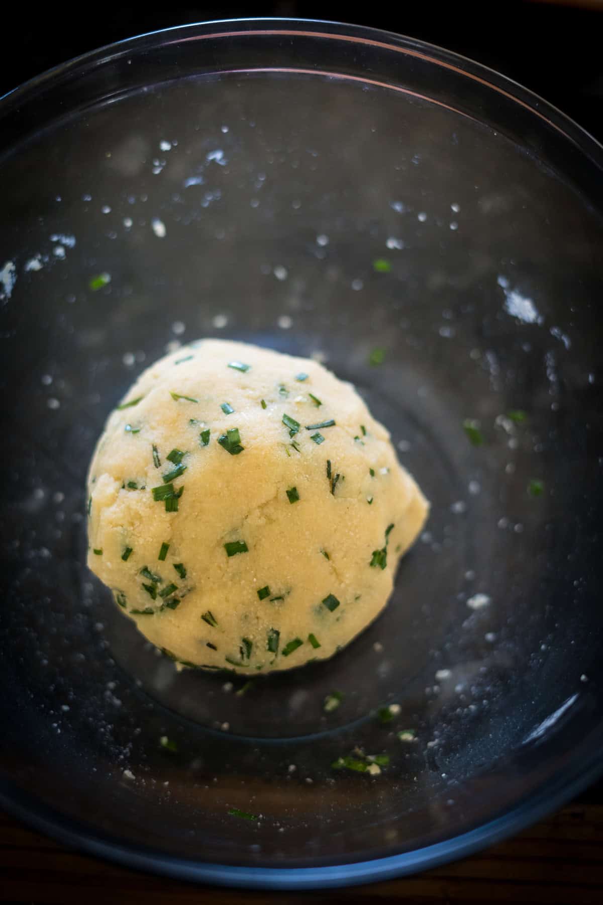 A ball of dough with green herbs mixed in, resting in a clear glass bowl, ready to be transformed into flavorful cheese crackers.