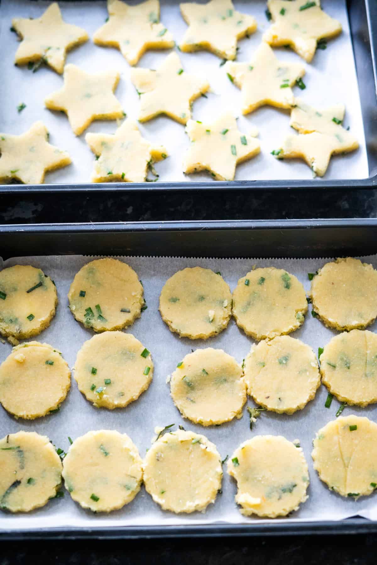 Two trays of unbaked cookies, resembling cheese crackers: one with star shapes and one with round shapes, both infused with green herbs, placed on parchment paper.